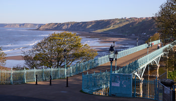 This landscape photograph shows the Scarborough cliffs and the ornate iron bridge stretching across the promenade in Yorkshire. The image was taken in the late afternoon during spring, with sunlight casting gentle shadows and illuminating the fresh green foliage of the trees. The cliffs of Scarborough dominate the background, where waves gently lap against the sandy shore and the open sea extends outwards. People can be seen walking along the bridge, which features decorative street lamps and intricate railings, reflecting classic Victorian architectural style. The location is a popular coastal destination in Yorkshire, and the photograph captures the scenic beauty and tranquil atmosphere of Scarborough’s shoreline and cliffs during the springtime.
